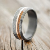 Shown here is "Castor", a custom, handcrafted men's wedding ring featuring a whiskey barrel oak and a 14K rose gold inlay on a hammered, fire-treated black zirconium band, upright facing left. Additional inlay options are available upon request.