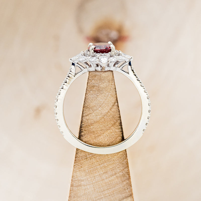 Shown here is "North Star", an oval garnet women's engagement ring with a diamond halo and accents, side view on stand. Many other center stone options are available upon request.