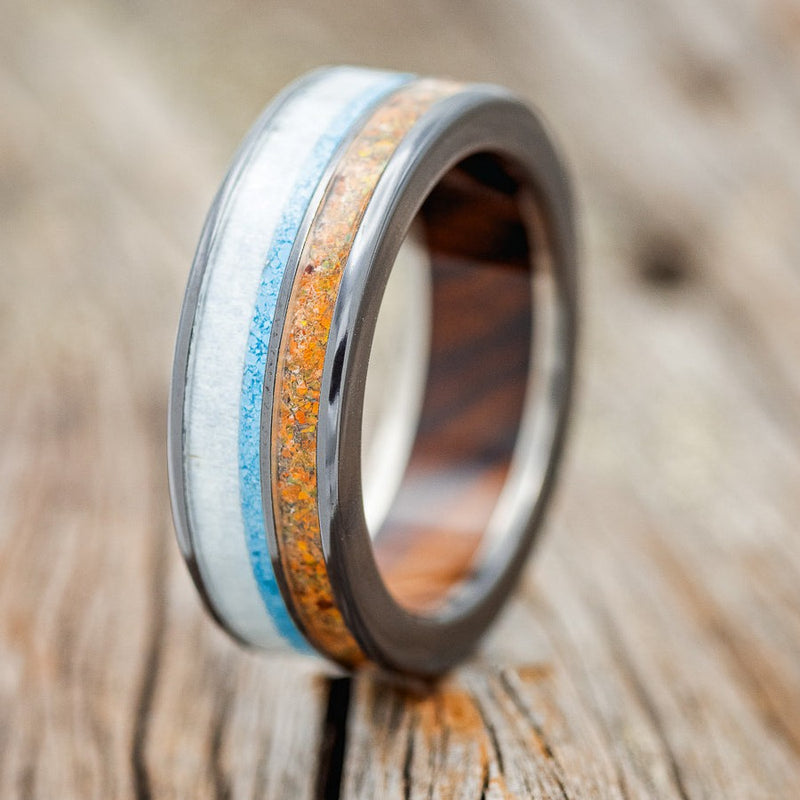 Shown here is "Element", a custom, handcrafted men's wedding ring featuring orange opal, elk antler, and hand-crushed turquoise on an ironwood lined, fire-treated, black zirconium band, upright facing left. Additional inlay options are available upon request.