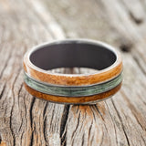 Shown here is "Glen", a custom, handcrafted men's wedding ring featuring whiskey barrel oak overlays and a fishing line inlay on a fire-treated black zirconium band, laying flat. Additional inlay options are available upon request.