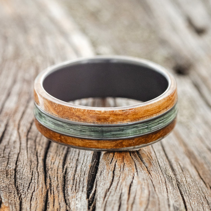 Shown here is "Glen", a custom, handcrafted men's wedding ring featuring whiskey barrel oak overlays and a fishing line inlay on a fire-treated black zirconium band, laying flat. Additional inlay options are available upon request.