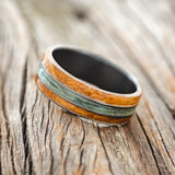 Shown here is "Glen", a custom, handcrafted men's wedding ring featuring whiskey barrel oak overlays and a fishing line inlay on a fire-treated black zirconium band, tilted left. Additional inlay options are available upon request.