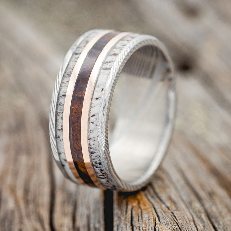 Shown here is "Rio", a custom, handcrafted men's wedding ring featuring antler and ironwood inlays divided by 2 14K rose gold inlays on an etched Damascus steel band, upright facing left. Additional inlay options are available upon request.