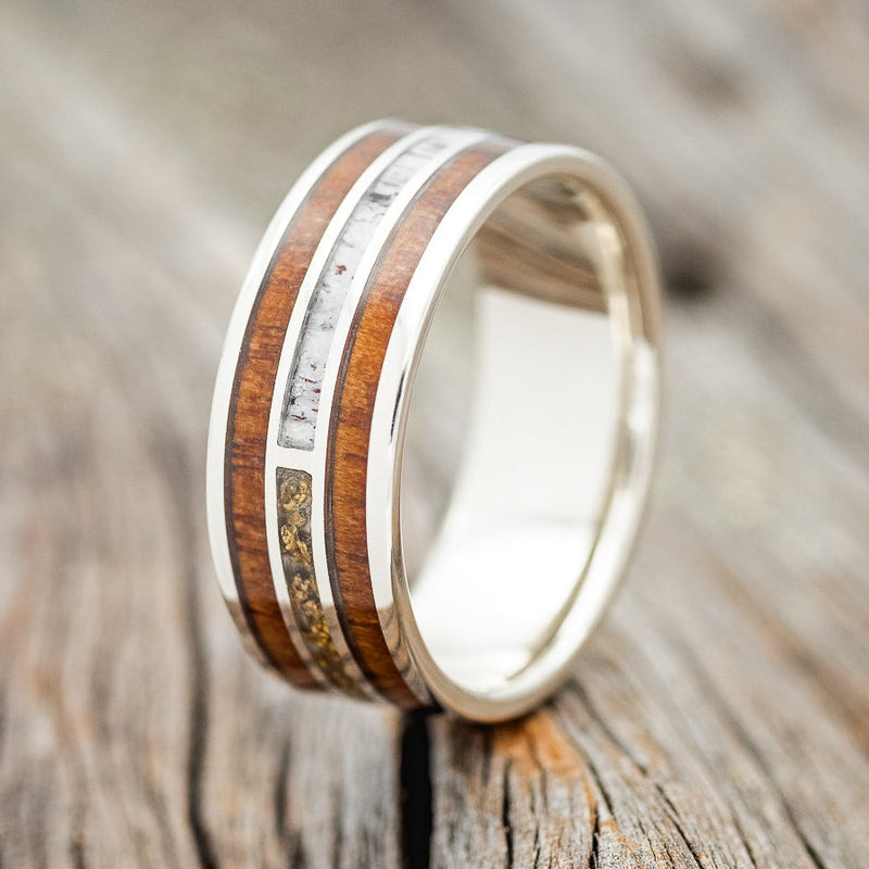 Shown here is "Ambrose", a custom, handcrafted men's wedding ring featuring Koa wood, antler, and gold nugget inlays on a silver band, upright facing left. Additional inlay options are available upon request.