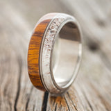 Shown here is "Banner", a custom, handcrafted men's wedding ring featuring ironwood and naturally shed antler with a guitar string inlay on a titanium band, upright facing left. Additional inlay options are available upon request.