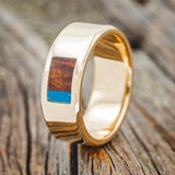 Shown here is a custom, handcrafted men's wedding ring featuring a rectangle inlay with redwood and turquoise on a 14K gold band, upright facing left. Additional inlay options are available upon request.