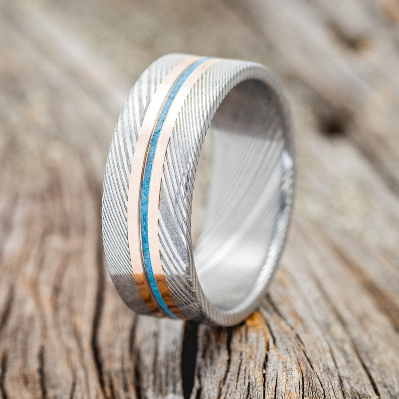 Shown here is "Nirvana", a custom, handcrafted men's wedding ring featuring a turquoise inlay and two 14K rose gold inlays on a Damascus steel band, upright facing left. Additional inlay options are available upon request.