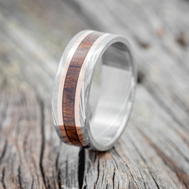 Shown here is "Kalder", a custom, handcrafted men's wedding ring featuring a koa wood inlay and two 1mm 14K rose gold inlays on a Damascus steel band, upright facing left. Additional inlay options are available upon request.