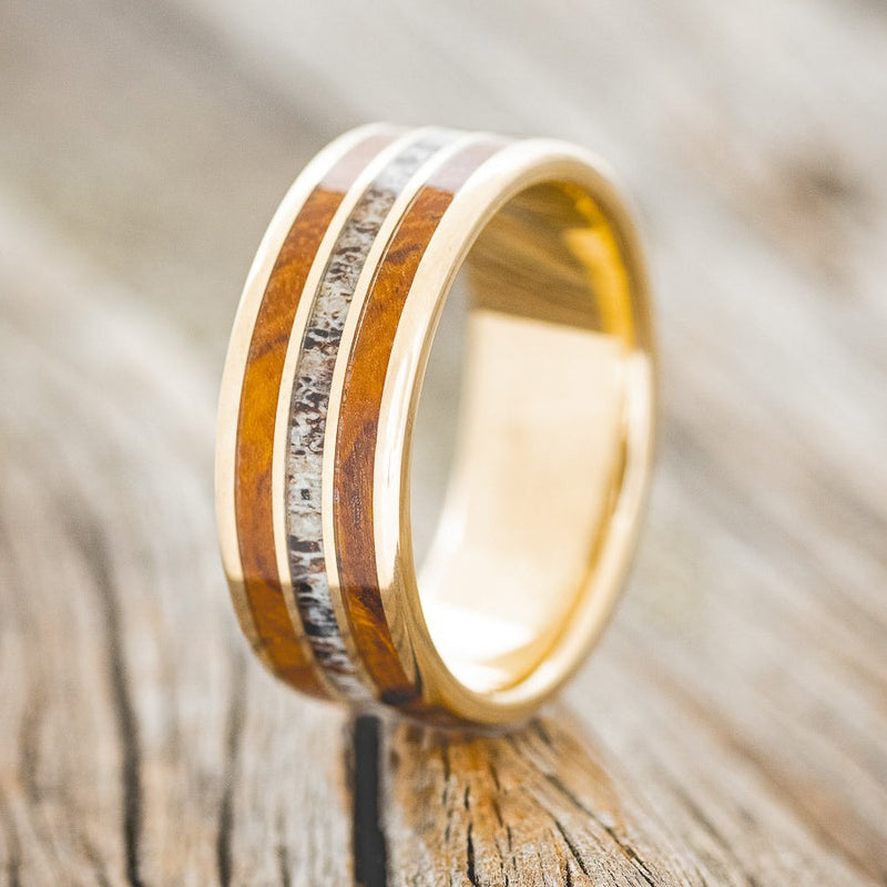 Shown here is "Rio", a custom, handcrafted men's wedding ring featuring 3 channels with redwood and antler inlays on a 14K gold band, upright facing left. Additional inlay options are available upon request.
