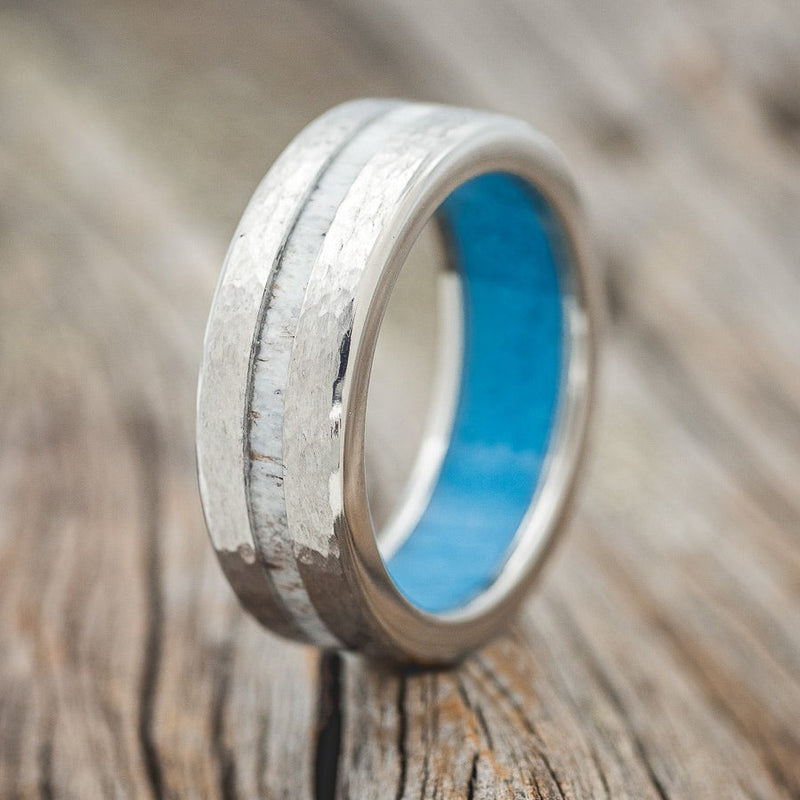 Shown here is "Vertigo", a custom, handcrafted men's wedding ring featuring a turquoise lining with an offset antler inlay on a hammered titanium band, upright facing left. Additional inlay options are available upon request.