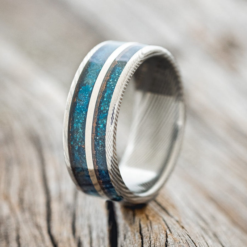 Shown here is "Raptor", a handcrafted men's wedding ring featuring two channels with patina copper inlays and a 14K white gold inlay, upright facing left. Additional inlay options are available upon request.