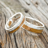 Shown here is "Raptor", a matching set of custom, handcrafted wedding rings featuring a mother of pearl and a whiskey barrel oak inlay, upright facing left. Additional inlay options are available upon request.