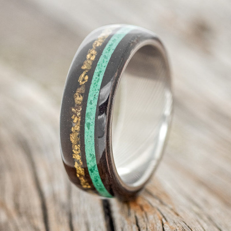 Shown here is "Remmy", a custom, handcrafted men's wedding ring featuring an African black wood overlay with gold nugget and malachite inlays on a Damascus steel band, upright facing left. Additional inlay options are available upon request.