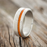 Shown here is "Vertigo", a custom, handcrafted men's wedding ring featuring an orange opal inlay on a Damascus steel band, upright facing left. Additional inlay options are available upon request.