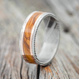 Shown here is "Hollis", a custom, handcrafted men's wedding ring featuring a Bethlehem olive wood inlay and two 1mm 14K rose gold inlays on a Damascus steel band, upright facing left. Additional inlay options are available upon request.