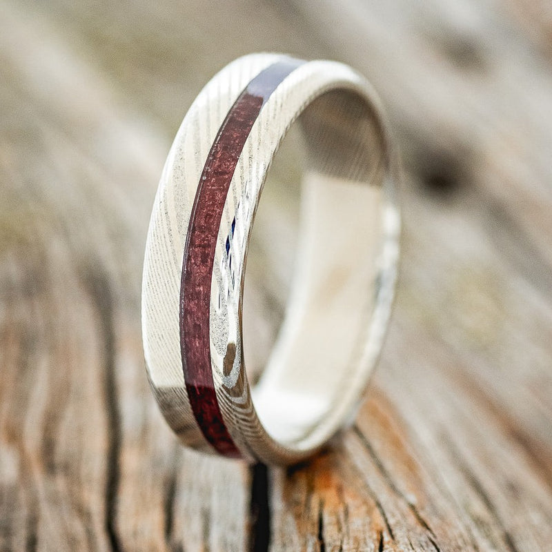 Shown here is "Vertigo", a custom, handcrafted men's wedding ring featuring an offset purpleheart wood inlay on a Damascus steel band, upright facing left. Additional inlay options are available upon request.