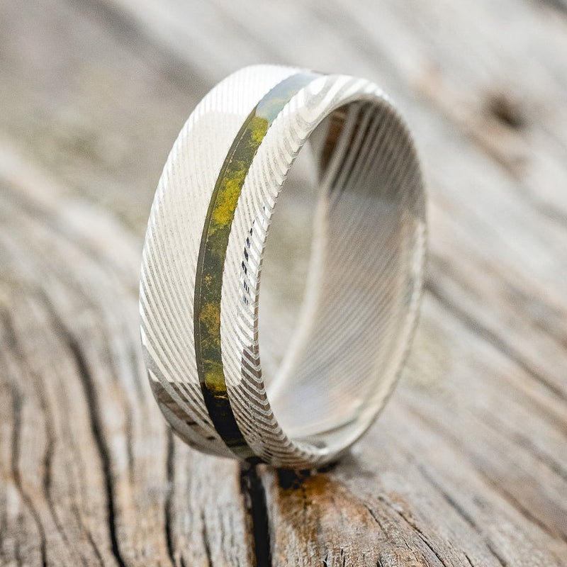 Shown here is "Vertigo", a custom, handcrafted men's wedding ring featuring an offset mossy patina copper inlay on a Damascus steel band, design shown facing right. Additional inlay options are available upon request.