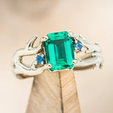 Shown here is "Artemis", an antler/branch-style lab-created emerald women's engagement ring with sapphire accents, on stand front facing. Many other center stone options are available upon request. 
