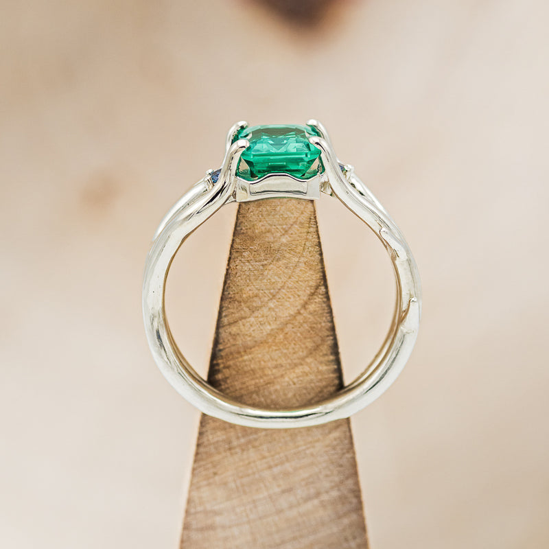 Shown here is "Artemis", an antler/branch-style lab-created emerald women's engagement ring with sapphire accents, side view on stand. Many other center stone options are available upon request.