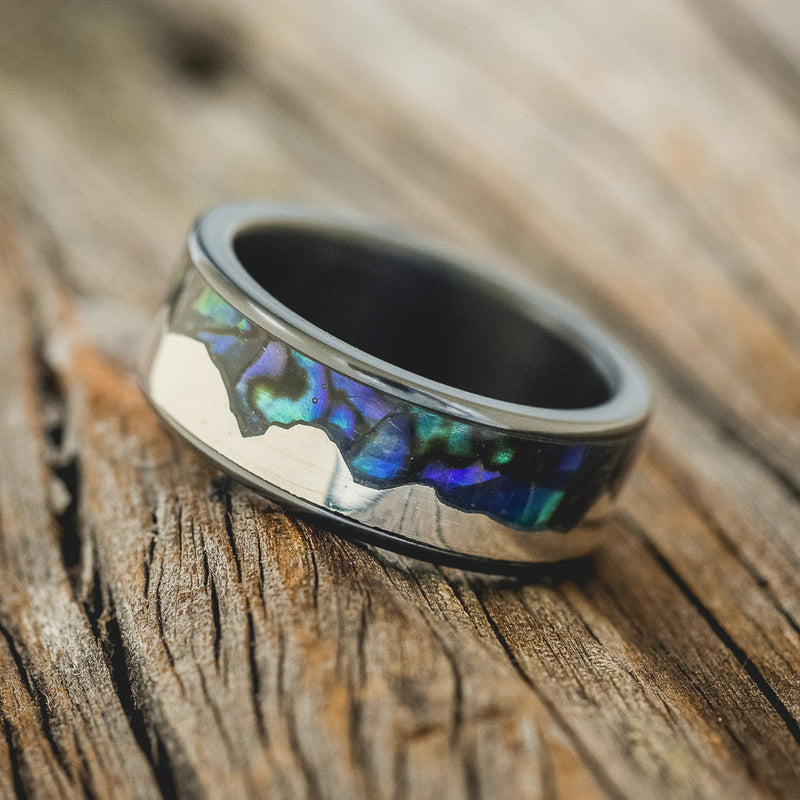Shown here is "Helios", a custom, handcrafted men's wedding ring featuring a mountain range using pieces of silver and paua shell inlay, tilted left. Additional inlay options are available upon request.
