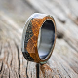 Shown here is "Ezra", a custom, handcrafted men's mountain wedding ring featuring a whiskey barrel oak overlay with jet stone and fire and ice opal mix, upright facing left. it is shown here on a fire-treated black zirconium band. Additional inlay options are available upon request.