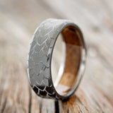 Shown here is "Echo", a handcrafted, custom embossed men's wedding ring featuring a dragon scale engraving with a whiskey barrel oak lining, upright facing left. This ring is pictured with a black zirconium band, giving it a higher contrast between embossment and engraving than other metal types will provide.