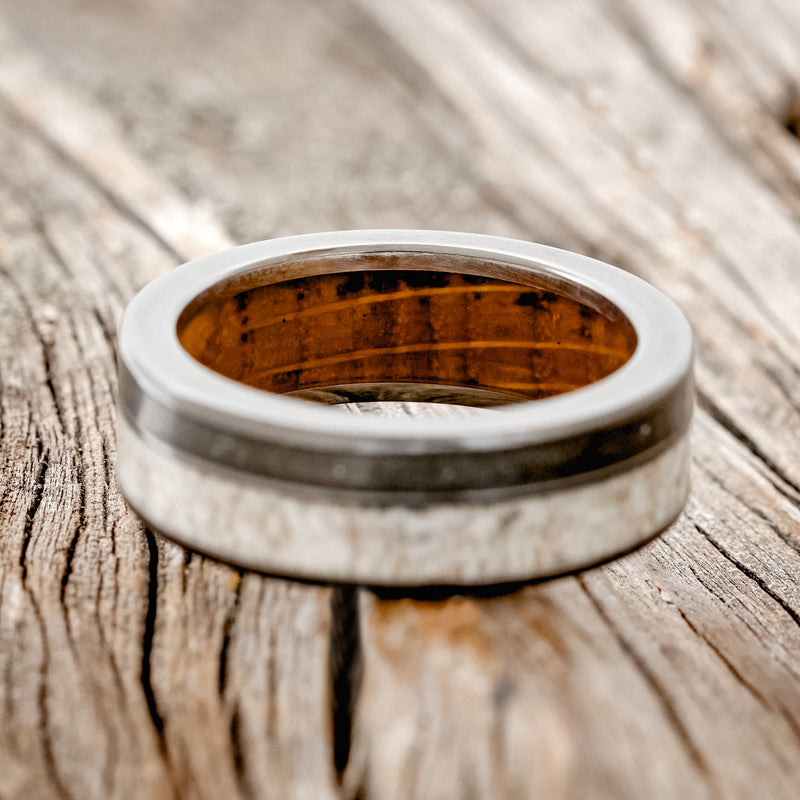 Shown here is "Raptor", a custom, handcrafted men's wedding ring featuring elk antler and iron ore inlays on a whiskey barrel oak lined black zirconium band, laying flat. Additional inlay options are available upon request.
