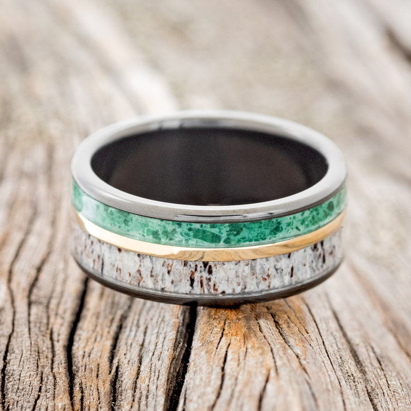 Shown here is "Raptor", a handcrafted men's wedding ring featuring two channels with malachite and antler inlays divided by a 14K yellow gold inlay, laying flat. Additional inlay options are available upon request.