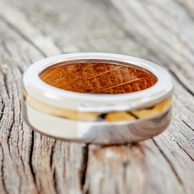 Shown here is "Vertigo", a handcrafted men's wedding ring featuring a spalted maple inlay on a whiskey barrel lined titanium band, laying flat. Additional inlay options are available upon request.
