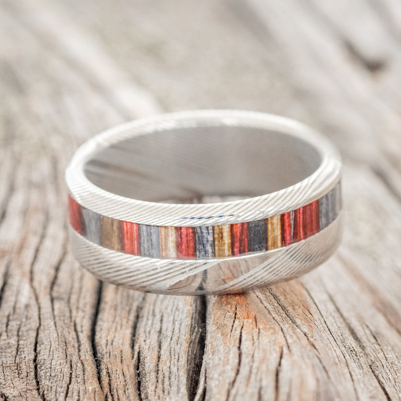 Shown here is "Castor", a custom, handcrafted men's wedding ring featuring a red, grey, and brown dyed birch wood inlay on a Damascus steel band, laying flat. Additional inlay options are available upon request.