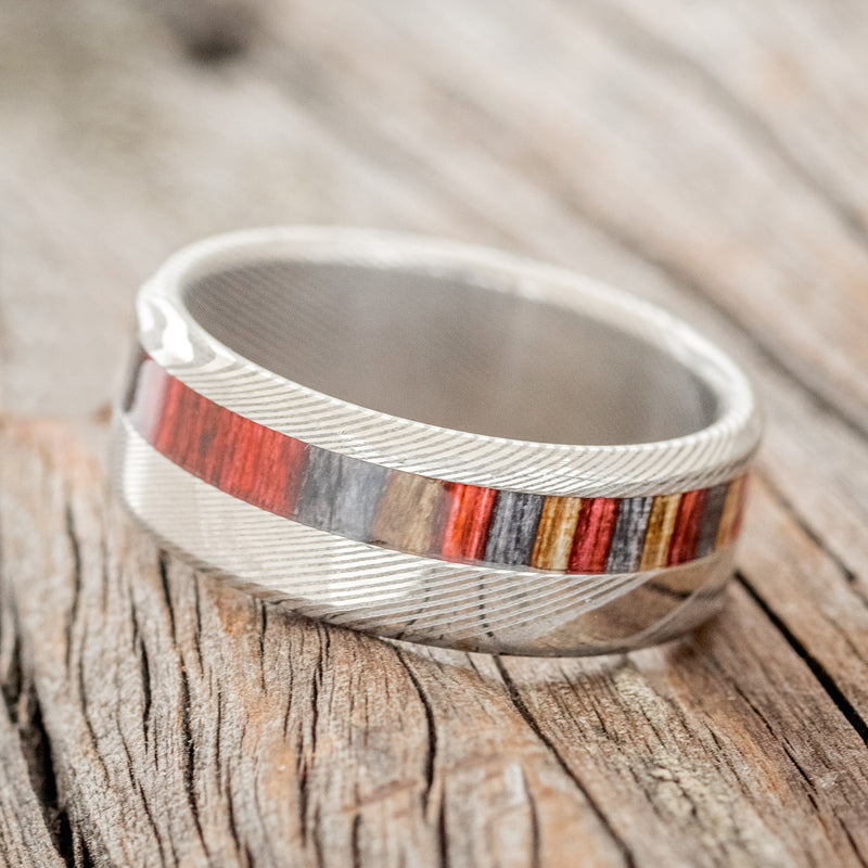 Shown here is "Castor", a custom, handcrafted men's wedding ring featuring a red, grey, and brown dyed birch wood inlay on a Damascus steel band, tilted left. Additional inlay options are available upon request.