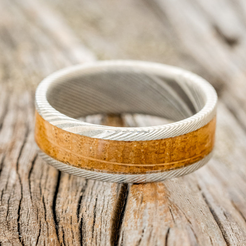 Shown here is "Kalder", a custom, handcrafted men's wedding ring featuring a whiskey barrel oak inlay on a Damascus steel band, laying flat. Additional inlay options are available upon request.