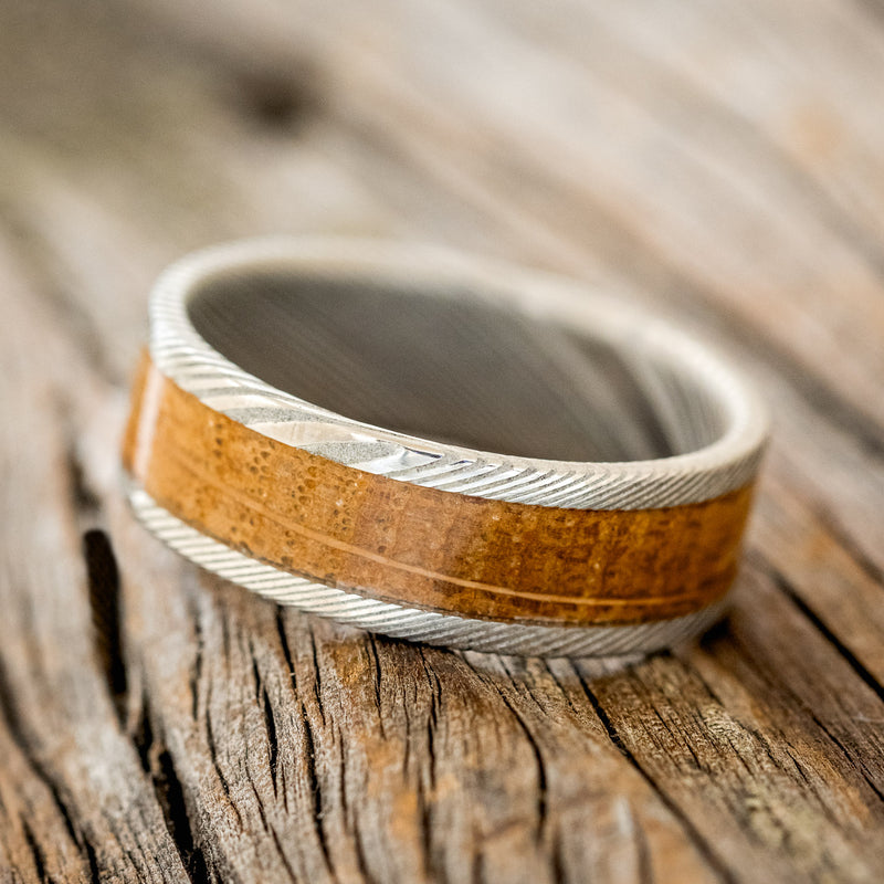 Shown here is "Kalder", a custom, handcrafted men's wedding ring featuring a whiskey barrel oak inlay on a Damascus steel band, tilted left. Additional inlay options are available upon request.