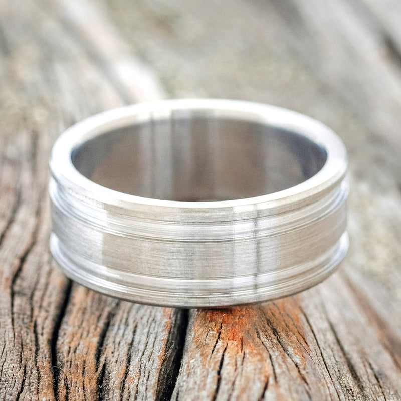 Shown here is "Ryder", a handcrafted men's wedding ring featuring a solid metal band that has two etched grooves and a brushed finish, laying flat.
