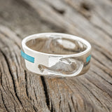 Shown here is "Hooked On You", a custom, handcrafted men's wedding ring featuring a fish hook shaped cut out and a turquoise inlay, laying flat. Additional inlay options are available upon request.