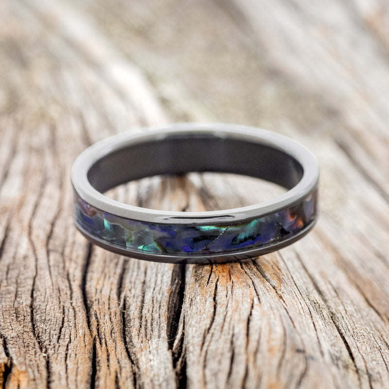 Shown here is "Perenna", a custom, handcrafted women's stacking band featuring a paua shell inlay on a fire-treated black zirconium band, laying flat. Additional inlay options are available upon request.
