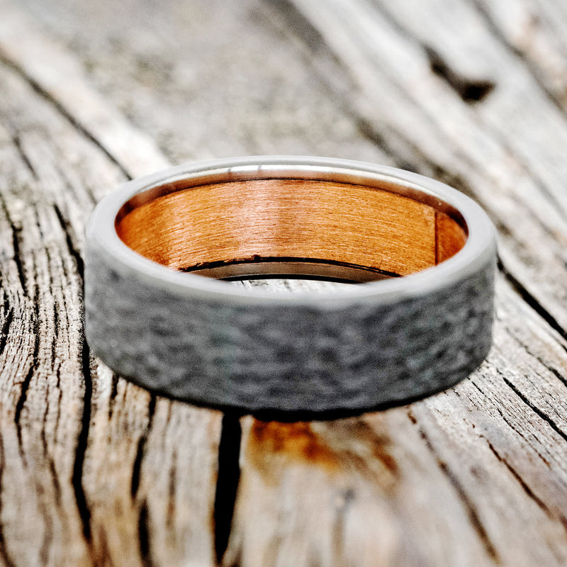 Shown here is a handcrafted men's wedding ring featuring a rustic copper lining with a hammered black zirconium band, laying flat. Additional lining options are available upon request.