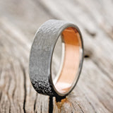 Shown here is a handcrafted men's wedding ring featuring a rustic copper lining with a hammered black zirconium band, upright facing left Additional lining options are available upon request.