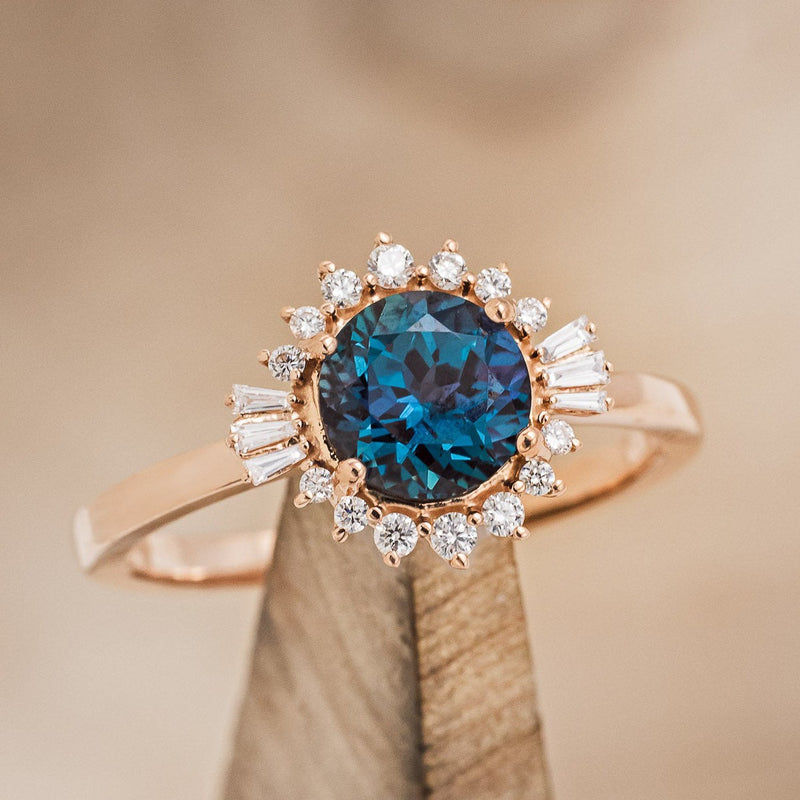 Shown here is "Coralie", a women's engagement ring with a lab-created alexandrite center stone with a starburst diamond halo, on stand front facing. Many other center stone options are available upon request.