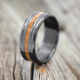 Shown here is "Austin", a custom, handcrafted men's wedding ring featuring a whiskey barrel stave oak inlay on a fire-treated black zirconium band, upright facing left. Additional inlay options are available upon request.