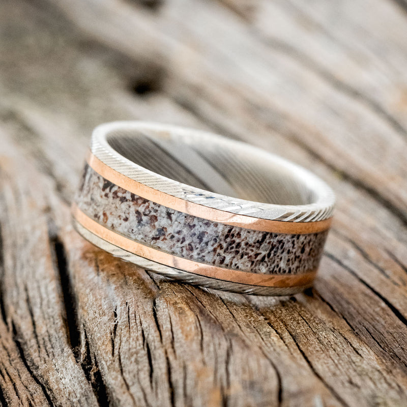Shown here is "Hollis", a custom, handcrafted men's wedding ring featuring an elk antler inlay and two 1mm 14K rose gold inlays on a Damascus steel band, tilted left. Additional inlay options are available upon request.