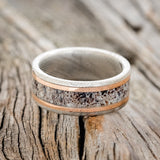 Shown here is "Hollis", a custom, handcrafted men's wedding ring featuring an elk antler inlay and two 1mm 14K rose gold inlays on a Damascus steel band, laying flat. Additional inlay options are available upon request.