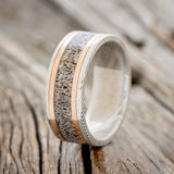 Shown here is "Hollis", a custom, handcrafted men's wedding ring featuring an elk antler inlay and two 1mm 14K rose gold inlays on a Damascus steel band, upright facing left. Additional inlay options are available upon request.