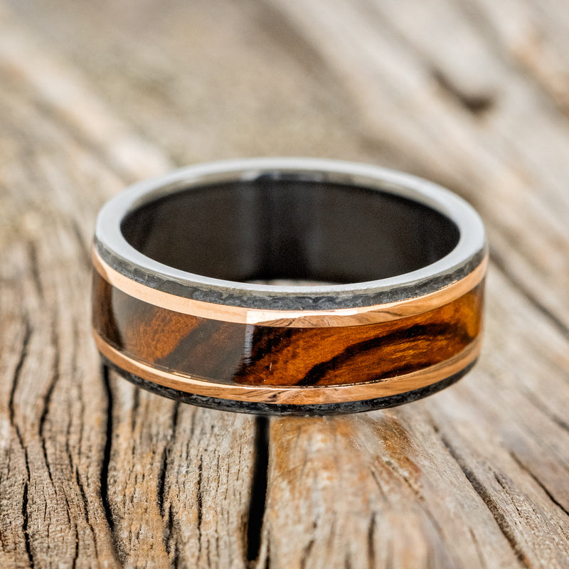 Shown here is "Hollis", a custom, handcrafted men's wedding ring featuring ironwood and 2 14K rose gold inlays with a hammered black zirconium band, laying flat. Additional inlay options are available upon request.