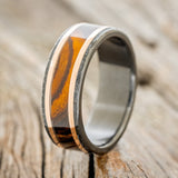 Shown here is "Hollis", a custom, handcrafted men's wedding ring featuring ironwood and 2 14K rose gold inlays with a hammered black zirconium band, upright facing left. Additional inlay options are available upon request.