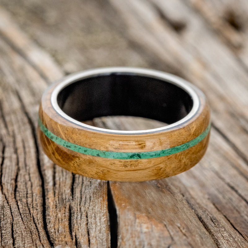 Shown here is "Remmy", a custom, handcrafted men's wedding ring featuring a whiskey barrel oak overlay and an offset malachite inlay on a fire-treated black zirconium band, laying flat. Additional inlay options are available upon request.