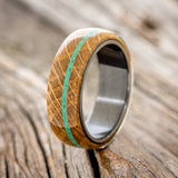 Shown here is "Remmy", a custom, handcrafted men's wedding ring featuring a whiskey barrel oak overlay and an offset malachite inlay on a fire-treated black zirconium band, upright facing left. Additional inlay options are available upon request.