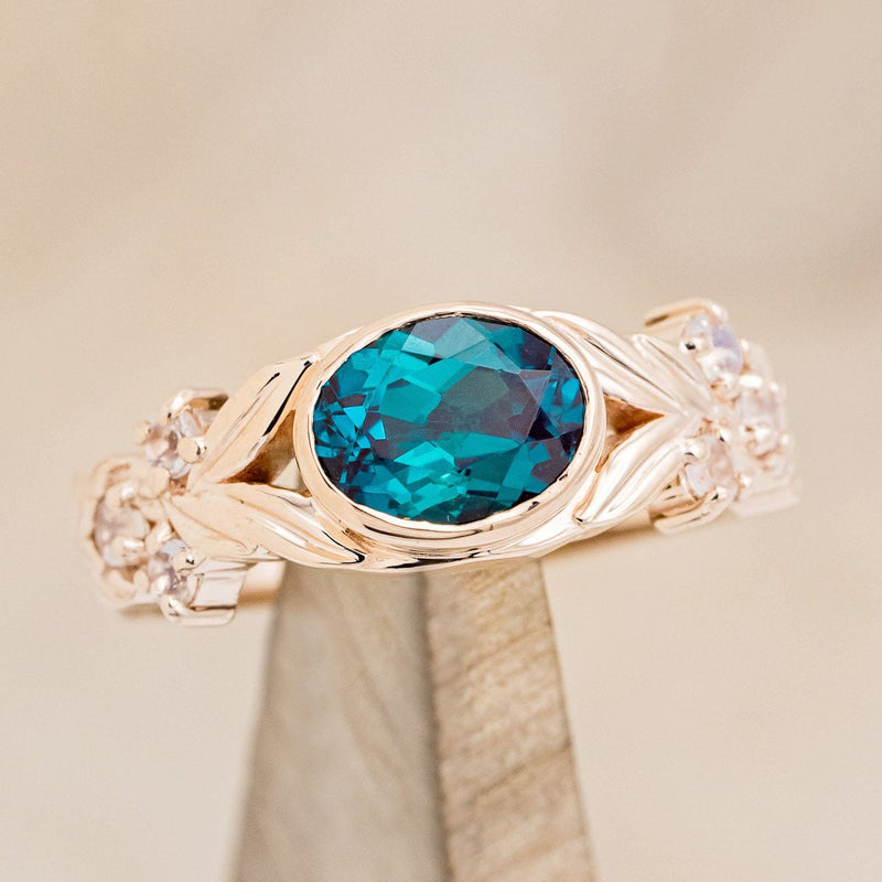 Shown here is "Elora", a vintage-style oval lab-created alexandrite women's engagement ring with floral details and moonstone accents, on stand tilted right. Many other center stone options are available upon request. 