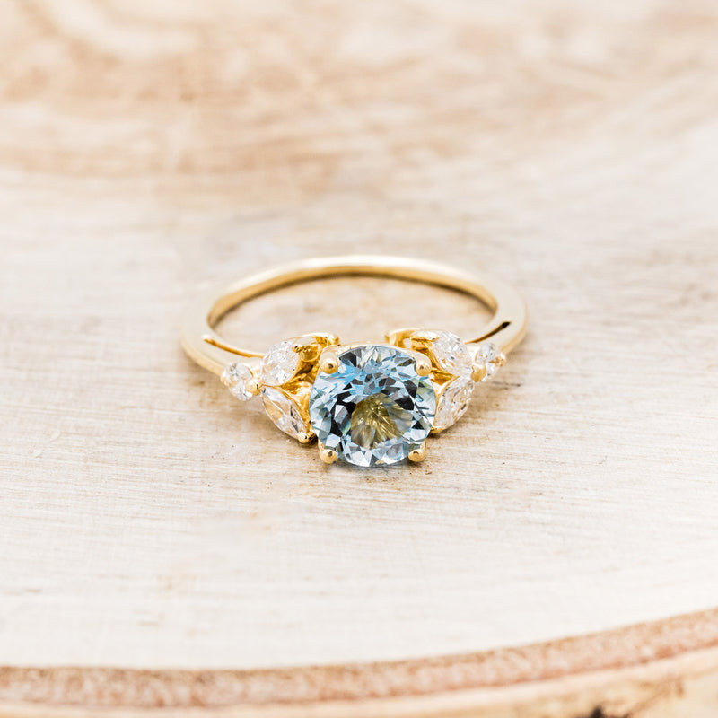 Shown here is "Blossom", a round cut aquamarine women's engagement ring with leaf-shaped diamond accents, front facing. Many other center stone options are available upon request.