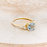 Shown here is "Blossom", a round cut aquamarine women's engagement ring with leaf-shaped diamond accents, facing right. Many other center stone options are available upon request.